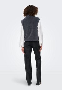 Personne aux cheveux bouclés portant une veste sans manches en polaire gris foncé, une chemise blanche à manches longues, un pantalon noir et des chaussures noires, vue de dos.
