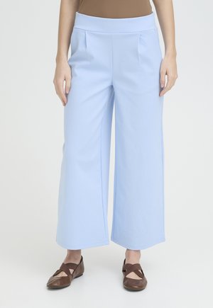 Femme portant un pantalon large bleu clair associé à des ballerines marron fermées avec des brides croisées, debout sur un fond blanc uni.