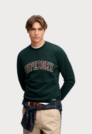 Young man with short brown hair wearing a dark green Superdry sweatshirt and beige pants with a plaid shirt tied around his waist.
