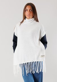 Poncho bianco lavorato a maglia con dolcevita alto, caratterizzato da un orlo frangiato e un'etichetta laterale. Indossato sopra un top a maniche lunghe scuro.