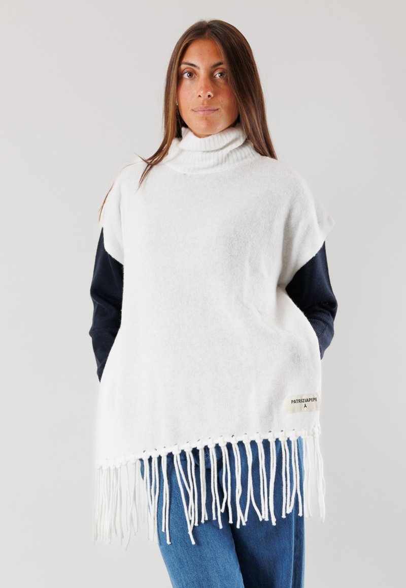 Poncho bianco lavorato a maglia con dolcevita alto, caratterizzato da un orlo frangiato e un'etichetta laterale. Indossato sopra un top a maniche lunghe scuro.