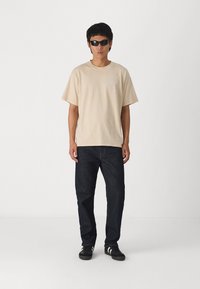Camiseta beige de manga corta, jeans azul oscuro y zapatillas negras. La figura lleva gafas de sol negras y está de pie contra un fondo blanco.