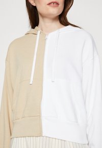 Felpa con zip crema e bianca, con un design a spacco, tasca a marsupio e lacci bianchi. Tessuto morbido con una vestibilità comoda.