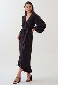 Robe noire à manches bouffantes, avec ourlet froncé et taille à nouer. Texture lisse, longueur genoux, assortie à des sandales à talons bloc noires.