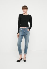 Črn, skrajšan gumbasti kardigan z ribasto teksturo in rahlo valovitim robom, v kombinaciji z svetlo modrimi hlačami visoke pasu in črnimi čevlji s peto.