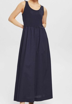 Femme portant une robe sans manches bleu marine, longue jusqu'aux chevilles, avec une taille froncée, debout les mains derrière le dos sur un fond clair uni.