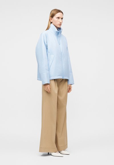 Γυναίκα που στέκεται με ανοιχτό γαλάζιο μπουφάν με ψηλό γιακά, μπεζ φαρδιά παντελόνια και λευκά μυτερά ψηλοτάκουνα, σε απλό φόντο.