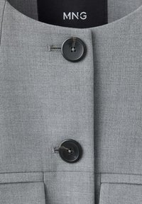 Gros plan d'une veste en tissu gris avec deux grands boutons foncés et l'étiquette de la marque "MNG" visible à l'intérieur du col.