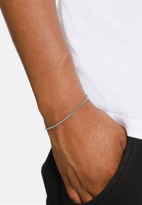 Zilveren gevlochten armband om een pols, met een gladde textuur, gecombineerd met een wit shirt en donkere broek. Hand in de zak geplaatst.