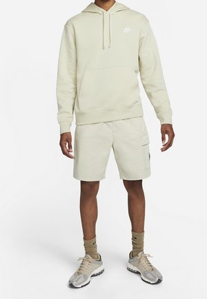 Modèle masculin portant un sweat à capuche Nike beige clair, un short cargo assorti, des chaussettes Nike beiges et des baskets gris-beige, debout sur un fond blanc.