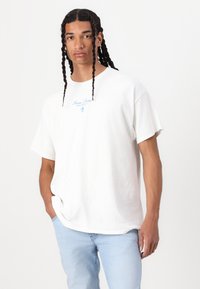 Witte katoenen t-shirt met korte mouwen, een ronde halslijn en blauwe tekstlogo boven een grafisch element op de voorkant. Draag het met lichtblauwe jeans.