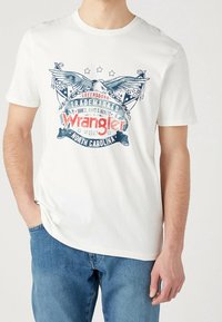 Bílý bavlněný tričko s barevným grafickým designem "Wrangler" s křídly a hvězdami. Kulatý výstřih a krátké rukávy, doplněné modrými džíny.