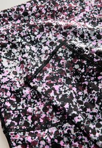 Gros plan sur un tissu noir avec des taches irrégulières roses, blanches et violet foncé, et une surpiqûre noire le long des bords.