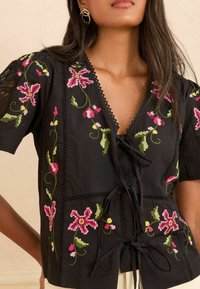 Melna izšūta blūze ar ziedu motīviem rozā un zaļā krāsā, V-veida kakla izgriezums ar saitēm, mežģīņu apdare uz piedurknēm un viegla kokvilnas auduma.