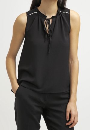 Blouse noire sans manches avec col à nouer sur le devant et fines bordures blanches sur les épaules, portée avec un pantalon noir.