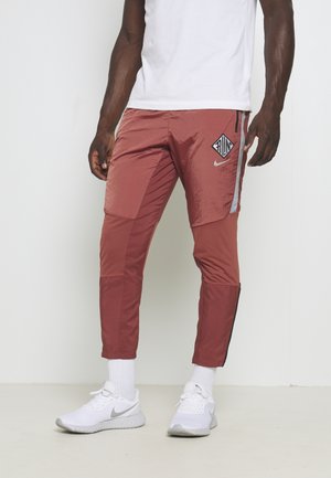 Man draagt roestkleurige hardloopbroek met "RUN" en Nike-logo's, witte sokken, witte Nike hardloopschoenen, staand tegen een witte achtergrond.