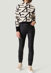 Gebreide trui in crèmekleur met zwarte abstracte lijnen, gecombineerd met nauwsluitende zwarte broek en beige sneakers, wat een contrast creëert in textuur en kleur.