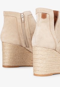 Botas de tobillo de ante beige con cremallera exterior y tacones de cuña de rafia trenzada. Parche de logo notable en la parte trasera.