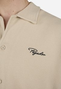 Beige strikket polo-skjorte med en teksturert overflate, med knappelukking og svart "Pegador"-logo brodert på brystet.
