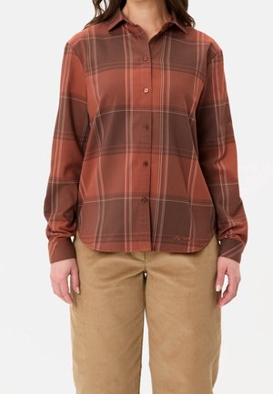 Kvinde iført en rust- og brunternet skorte med lange ærmer og beige kordfløjlsbukser, stående mod en ensfarvet baggrund.