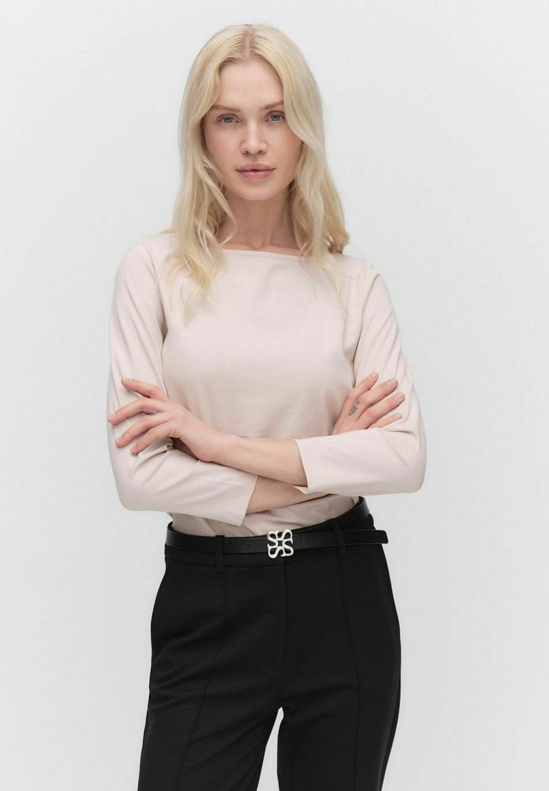 Helles rosa Langarmoberteil, kombiniert mit schwarzen Hosen. Das Outfit zeichnet sich durch ein elegantes Design und einen silbernen Akzent am Gürtel aus. Glatte Textur.
