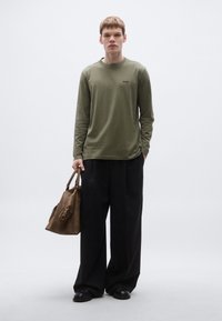 Μακρυμάνικο t-shirt σε ελιά πράσινο με μικρό λογότυπο, συνδυασμένο με φαρδιά μαύρη παντελόνια και καφέ δερμάτινη τσάντα. Λεία υφή, άνετη εφαρμογή.