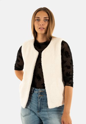 Gilet sans manches couleur crème en tissu doux et texturé, avec une ouverture à l'avant et sans fermetures, porté sur un haut noir à fleurs.