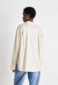 Beige linen shirt with a back pleat, a pointed collar, and buttoned cuffs, paired with light blue denim pants.