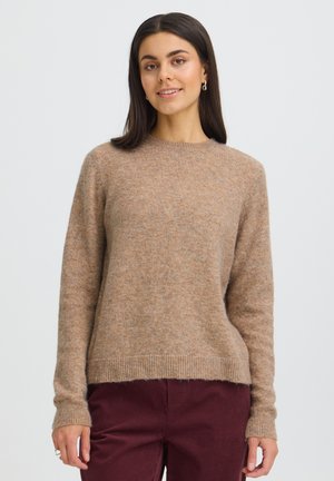 Femme aux longs cheveux foncés porte un pull en maille brun clair et un pantalon bordeaux, debout devant un fond clair uni, face à l'avant.