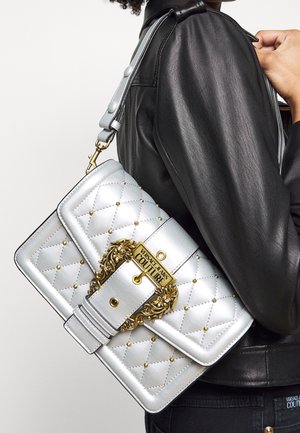Zilveren gewatteerde handtas met gouden hardware en studs, met een opvallend logo aan de voorkant. De riem is verstelbaar en gedetailleerd.