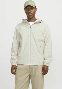 Jachetă zip-up de un gri deschis, cu glugă, manșete elastice și buzunare laterale, asortată cu o șapcă corespunzătoare și pantaloni bej deschis.