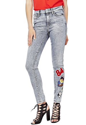 Frau trägt hellgraue Skinny-Jeans mit Acid-Waschung und bunten Aufnähern am Unterschenkel, schwarze Schnür-Pumps mit Absatz und ein rotes Oberteil.