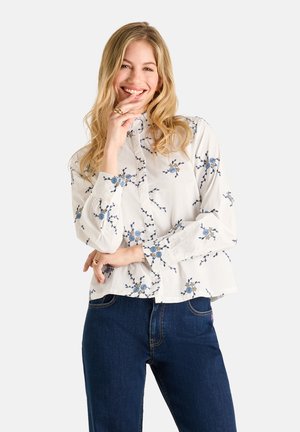 Frau lächelt mit langen blonden Haaren, trägt eine weiße geblümte Bluse und dunkelblaue Jeans, posiert mit der Hand nahe am Gesicht.