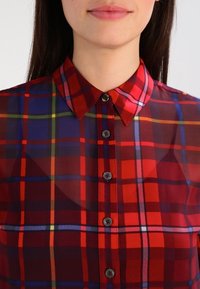 Camicia a quadri rossa e blu con colletto classico, caratterizzata da bottoni grigi e una texture di tessuto leggero e trasparente.