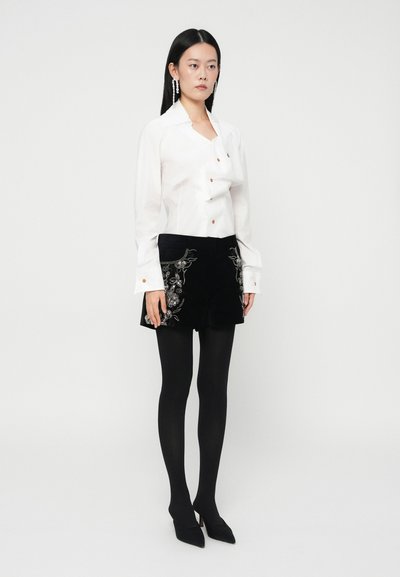 Chemise blanche boutonnée avec un design plié unique, associée à un short en velours noir orné de broderies florales. Collants noirs et talons pointus.