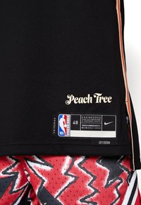 Camiseta de baloncesto negra con "Peach Tree" en dorado en cursiva. Presenta una etiqueta con la talla 48 y la marca Nike. Pantalones cortos rojos y negros con un patrón en la parte inferior.