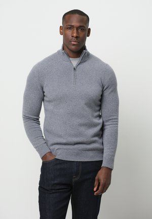 Maglioncino grigio con zip al collo, realizzato in materiale morbido, design aderente con maniche lunghe, polsini e orlo a costine, indossato sopra una camicia chiara e jeans scuri.
