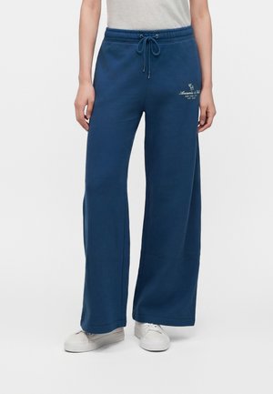 Personne portant un pantalon de survêtement bleu à jambes larges avec une taille à cordon et des baskets blanches, debout sur un fond neutre.