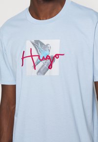 Blue t-shirt with a graphic of a blue budgerigar on a skateboard and red cursive "Hugo" text across the chest.
