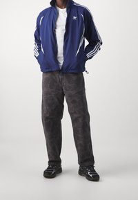 Blue sports jacket with white stripes, paired with dark patterned pants and black sneakers. Jacket features a zip and logo on the chest.