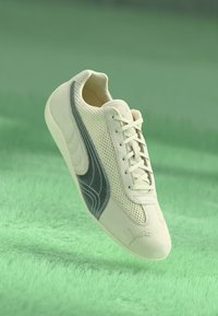 Hvid sneaker med sort Puma-logo og mesh-detaljer svævende over en grøn tekstureret overflade.