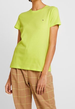 Person iført en knaldgrøn kortærmet T-shirt med et lille logo, kombineret med brune ternede bukser med gule linjer.