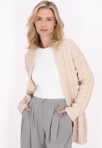 Beige kötött kardigán, kábelmintával, hosszú ujjakkal és nyitott elülső résszel. Fehér blúz és szürke pliszírozott nadrág fölött viselve.
