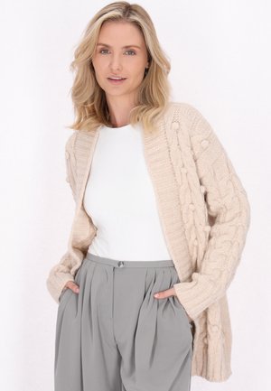 Beige pletený kardigan s copánkovým vzorem, dlouhými rukávy a otevřeným předním dílem. Nositý přes bílé tričko a šedé skládací kalhoty.