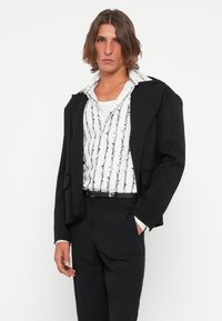 Schwarzer maßgeschneiderter Blazer über einem weißen Hemd mit einem schwarzen Bambusmuster; das Model trägt schwarze Hose, die einen modernen Schnitt und einen geschichteten Look zeigt.