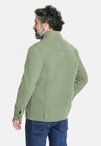 Hombre con cabello rizado y barba, vistiendo una chaqueta verde claro y jeans azules, de perfil con la cabeza girada hacia la izquierda.