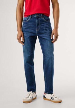 Homme portant un jean slim bleu, des baskets blanches avec des détails noirs et rouges, et une chemise rouge sur un fond clair uni.