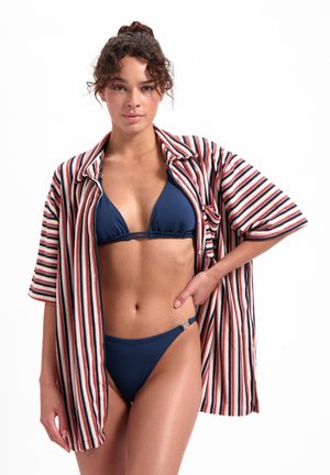 Camicia a righe rossa, blu navy e bianca con maniche corte sopra un bikini triangolare blu navy. Il bikini presenta un accento a forma di anello unico sul lato.