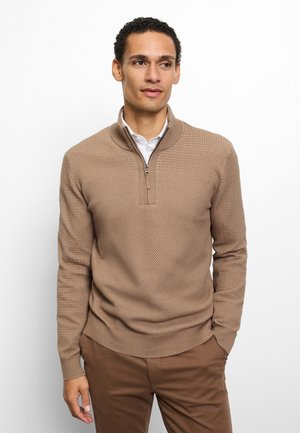 Jonge man die een beige gestructureerde trui met kwart-ritssluiting draagt over een wit overhemd en een bruine broek, staand tegen een witte achtergrond.