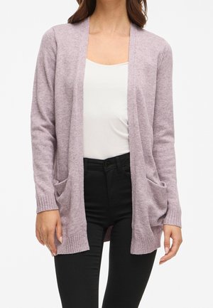 Femme portant un cardigan violet pâle ouvert sur le devant avec des poches, un haut blanc et un pantalon noir, debout devant un fond blanc.